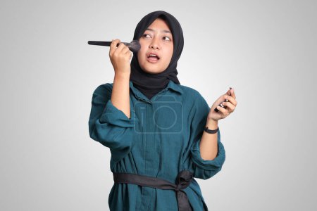 Téléchargez les photos : Portrait de femme musulmane asiatique excitée avec hijab, appliquer la poudre de fond de teint ou rougir avec un pinceau de maquillage sur sa joue. Image isolée sur fond blanc - en image libre de droit