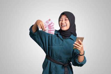Téléchargez les photos : Portrait de femme hijab asiatique excitée en tenue décontractée montrant cent mille roupies tout en tenant un téléphone portable. Concept financier et d'épargne. Image isolée sur fond blanc - en image libre de droit