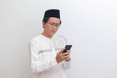 Téléchargez les photos : Portrait de jeune homme musulman asiatique tenant un téléphone portable avec une expression souriante sur le visage. Image isolée sur fond blanc - en image libre de droit