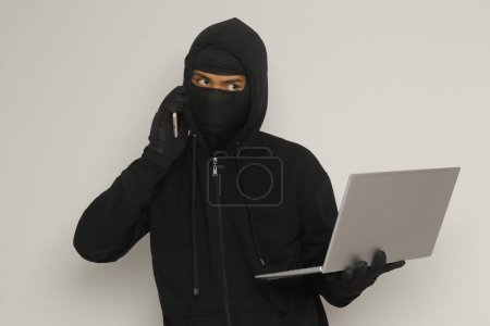 Téléchargez les photos : Portrait d'un homme mystérieux portant un sweat à capuche noir et un masque faisant de l'activité de piratage sur un ordinateur portable, un pirate tenant un téléphone portable. Concept de cybersécurité. Image isolée sur fond gris - en image libre de droit