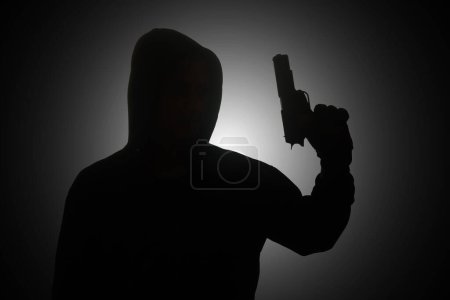 Téléchargez les photos : Homme mystérieux portant un sweat à capuche noir tenant un pistolet, tirant avec un pistolet. Silhouette et image concept sombre - en image libre de droit