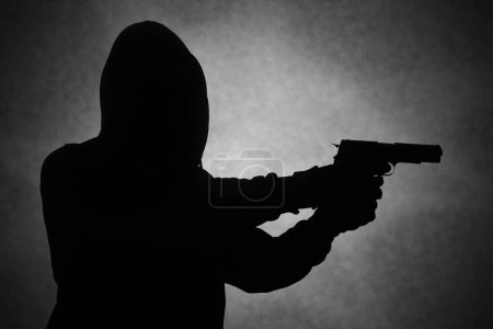 Téléchargez les photos : Homme mystérieux portant un sweat à capuche noir tenant un pistolet, tirant avec un pistolet. Silhouette et image concept sombre - en image libre de droit