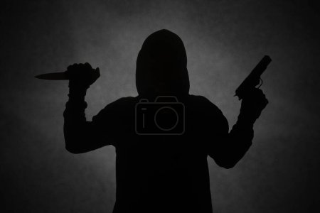 Téléchargez les photos : Homme mystérieux portant un sweat à capuche noir tenant un pistolet, tirant avec un pistolet tout en tenant un couteau. Silhouette et image concept sombre - en image libre de droit