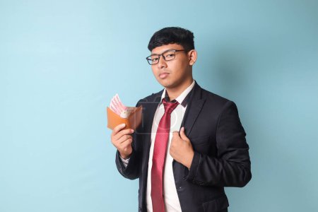 Téléchargez les photos : Portrait de jeune homme d'affaires asiatique en costume décontracté avec des expressions de confiance fixant le collier et tenant portefeuille en cuir avec mille roupies. Image isolée sur fond bleu - en image libre de droit