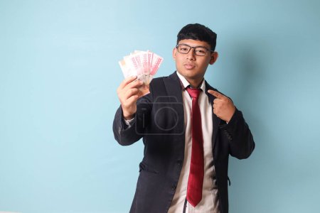 Téléchargez les photos : Portrait de jeune homme d'affaires asiatique en costume décontracté pointant vers mille roupies avec des expressions sérieuses. Image isolée sur fond bleu - en image libre de droit