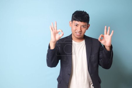Téléchargez les photos : Portrait de jeune homme d'affaires asiatique en costume décontracté avec des expressions heureuses et des gestes de main double accord. Image isolée sur fond bleu - en image libre de droit