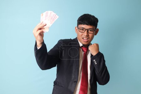 Téléchargez les photos : Portrait de jeune homme d'affaires asiatique en costume casual. avec des expressions heureuses tenant mille roupies et célébrant. Image isolée sur fond bleu - en image libre de droit