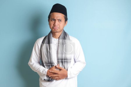 Téléchargez les photos : Portrait d'un jeune homme musulman asiatique qui ressent de la douleur dans son estomac. Concept de mal d'estomac. Image isolée sur fond bleu - en image libre de droit