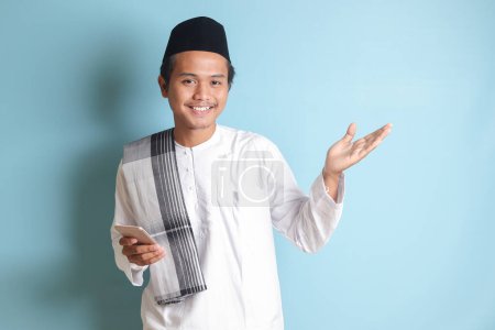 Téléchargez les photos : Portrait de jeune homme musulman asiatique tenant un téléphone portable avec une expression souriante sur le visage tout en pointant du doigt vers le côté. Image isolée sur fond bleu - en image libre de droit