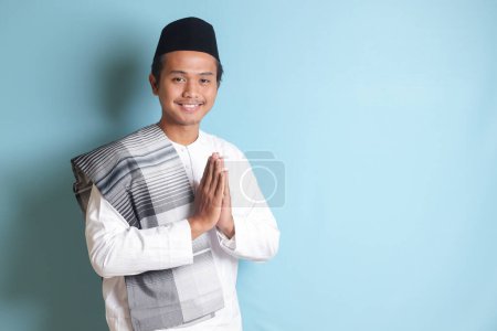 Téléchargez les photos : Portrait d'homme musulman asiatique en chemise koko blanche avec bonnet de crâne montrant des excuses et geste de bienvenue. Image isolée sur fond bleu - en image libre de droit