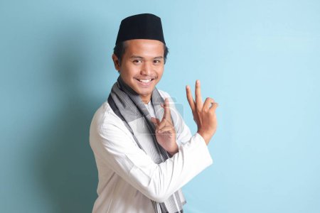 Téléchargez les photos : Portrait d'homme musulman asiatique en chemise koko blanche faisant un geste d'arts martiaux ou un silat de crayon. Image isolée sur fond bleu - en image libre de droit