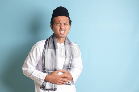 Téléchargez les photos : Portrait d'un jeune homme musulman asiatique qui ressent de la douleur dans son estomac. Concept de mal d'estomac. Image isolée sur fond bleu - en image libre de droit