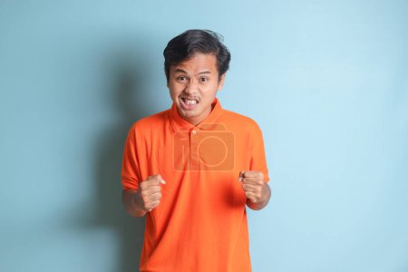 Téléchargez les photos : Homme asiatique ennuyé en chemise orange faisant geste de la main en colère avec les doigts. Image isolée sur fond bleu - en image libre de droit