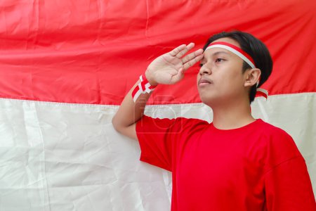 Téléchargez les photos : Portrait d'homme asiatique attrayant en chemise rouge avec ruban rouge et blanc sur la tête, donnant une pose de salut avec la main devant le drapeau indonésien - en image libre de droit