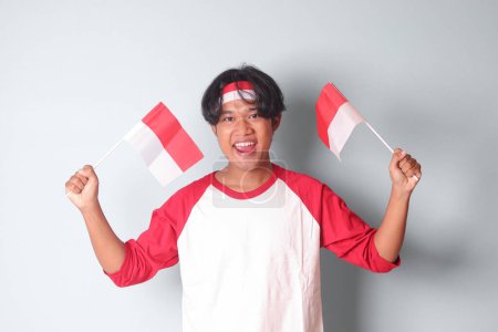 Photo for Portrait of attractive Asian man in t-shirt with red and white ribbon on head, holding indonesia flag while raising his fist, celebrating success. Isolated image on gray background - Royalty Free Image