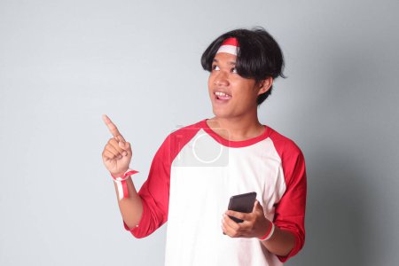 Téléchargez les photos : Portrait d'homme asiatique attrayant en t-shirt avec ruban rouge et blanc sur la tête, tenant le téléphone portable tout en levant le drapeau indonésien et pointant du doigt. Image isolée sur fond gris - en image libre de droit