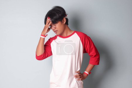 Téléchargez les photos : Portrait d'homme asiatique séduisant en t-shirt avec un ruban rouge et blanc sur la tête, ayant une migraine, touchant son temple. Concept des maux de tête. Image isolée sur fond gris - en image libre de droit