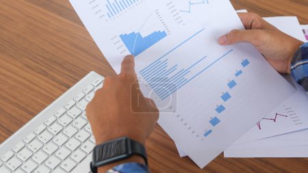 Primer plano de la mano de trabajador independiente masculino que trabaja en el ordenador portátil y señala con el dedo los datos de análisis con el gráfico de negocios y el diagrama de información en el escritorio de la oficina por la mañana