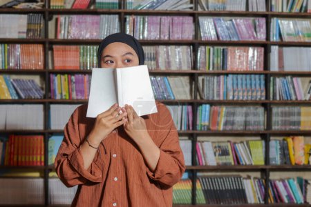 Téléchargez les photos : Portrait de femme hijab asiatique couvrant sa demi-tête d'un livre devant la bibliothèque. Une musulmane lisant un livre. Notion d'alphabétisation et de connaissance - en image libre de droit