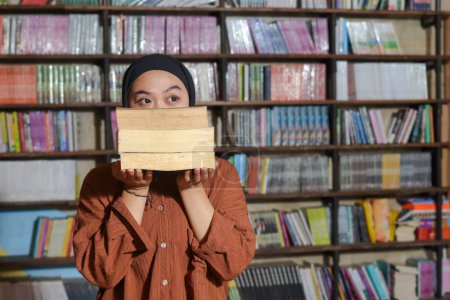 Téléchargez les photos : Portrait de femme hijab asiatique couvrant sa tête avec une pile de livres devant l'étagère de la bibliothèque. Une musulmane lisant un livre. Notion d'alphabétisation et de connaissance - en image libre de droit