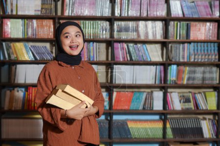 Téléchargez les photos : Portrait d'une femme hijab asiatique tenant un livre devant une bibliothèque. Une musulmane lisant un livre. Notion d'alphabétisation et de connaissance - en image libre de droit