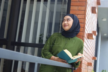 Téléchargez les photos : Portrait de femme hijab asiatique attrayant tenant livre dans le balcon. Une musulmane lisant un livre. Notion d'alphabétisation et de connaissance - en image libre de droit