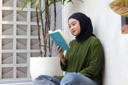 Téléchargez les photos : Portrait de femme hijab asiatique attrayant livre de lecture dans le balcon. Fille musulmane jouissant de temps libre. Notion d'alphabétisation et de connaissance - en image libre de droit