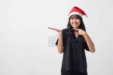 Téléchargez les photos : Portrait de femme asiatique attrayante avec chapeau rouge Santa se sentant heureux, pointant et montrant le produit sur le côté avec le doigt. Nouvel an et concept de Noël. Image isolée sur fond blanc - en image libre de droit