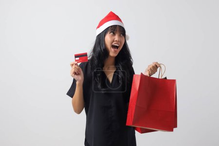 Téléchargez les photos : Portrait de femme asiatique attrayante avec chapeau rouge Santa montrant carte de crédit tout en tenant des sacs à provisions. Paiement sans espèces. Nouvel an et concept de Noël. Image isolée sur fond blanc - en image libre de droit