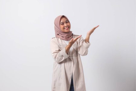 Téléchargez les photos : Portrait de femme asiatique excitée hijab en costume décontracté pointant et montrant le produit dans son côté avec le doigt. Concept de femme d'affaires. Image isolée sur fond blanc - en image libre de droit