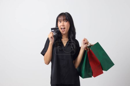 Téléchargez les photos : Portrait de femme asiatique excitée en tenant des sacs à provisions en papier coloré et montrant la carte de crédit. Fille Shopaholic et concept d'achat discount. Image isolée sur fond blanc - en image libre de droit