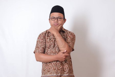 Téléchargez les photos : Portrait d'homme asiatique confus portant chemise batik et chanson debout sur fond gris, réfléchissant à la question avec la main sur le menton - en image libre de droit