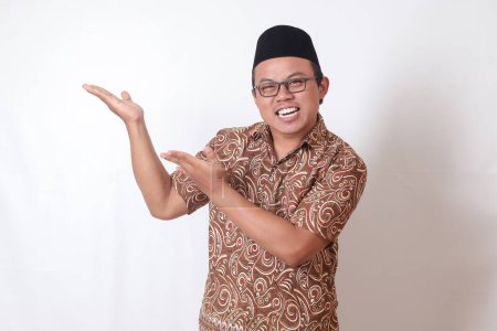 Téléchargez les photos : Portrait d'homme asiatique excité portant une chemise batik et chantant souriant et regardant la caméra pointant avec deux mains et doigts sur le côté. Image isolée sur fond gris - en image libre de droit