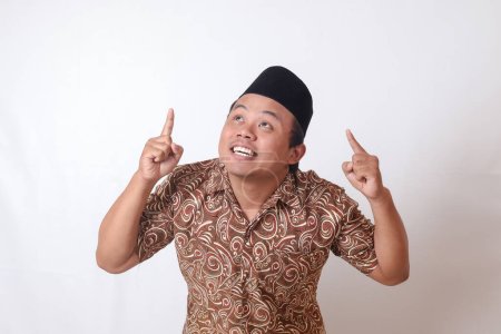 Téléchargez les photos : Portrait d'homme asiatique excité portant une chemise batik et chantant souriant et regardant la caméra pointant avec deux mains et les doigts vers le haut. Image isolée sur fond gris - en image libre de droit