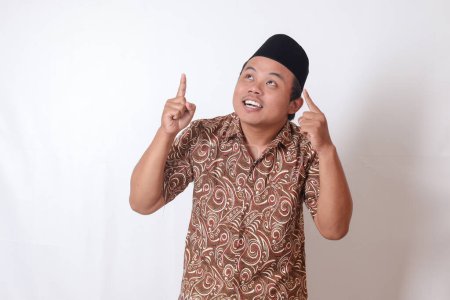 Téléchargez les photos : Portrait d'homme asiatique excité portant une chemise batik et chantant souriant et regardant la caméra pointant avec deux mains et les doigts vers le haut. Image isolée sur fond gris - en image libre de droit