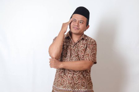 Téléchargez les photos : Portrait d'un homme asiatique souffrant portant une chemise en batik et chantant ayant mal à la tête tout en touchant son front. Image isolée sur fond gris - en image libre de droit