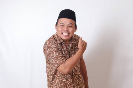 Téléchargez les photos : Portrait d'homme asiatique excité portant une chemise batik et chantant souriant et regardant la caméra, faisant les pouces vers le haut geste de la main. Image isolée sur fond gris - en image libre de droit