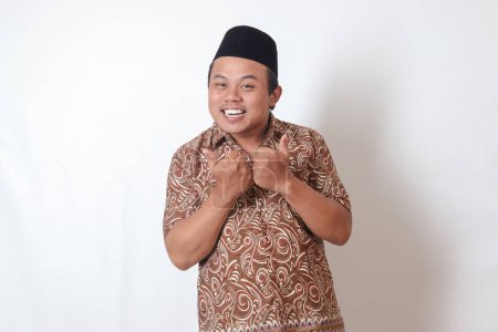 Photo for Portrait of excited Asian man wearing batik shirt and songkok smiling and looking at camera, making thumbs up hand gesture. Isolated image on gray background - Royalty Free Image