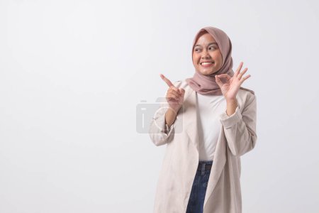 Téléchargez les photos : Portrait de femme hijab asiatique excitée en costume décontracté montrant ok geste de la main et souriant en regardant la caméra. Concept publicitaire. Image isolée sur fond blanc - en image libre de droit