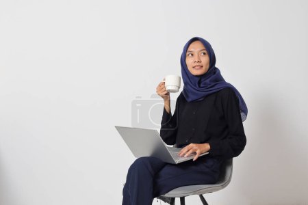 Téléchargez les photos : Portrait de femme hijab asiatique excitée en chemise décontractée assise sur une chaise, travaillant sur un ordinateur portable tout en buvant une tasse de café. Concept de femme d'affaires. Image isolée sur fond blanc - en image libre de droit