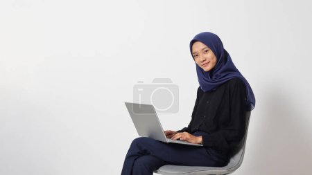 Téléchargez les photos : Portrait de femme hijab asiatique excitée en chemise décontractée assise sur une chaise, travaillant sur un ordinateur portable. Un employé ou un étudiant qui fait sa tâche et son travail. Concept de femme d'affaires. Image isolée sur fond blanc - en image libre de droit
