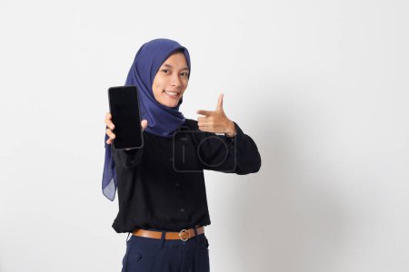 Téléchargez les photos : Portrait de femme asiatique excitée hijab en chemise décontractée montrant écran vierge téléphone portable maquette tout en pointant et en présentant le produit. Image isolée sur fond blanc - en image libre de droit