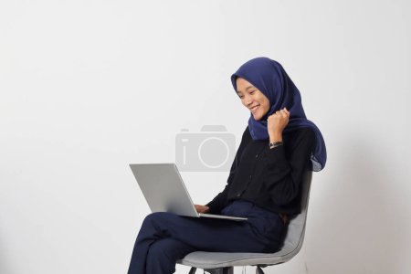 Téléchargez les photos : Portrait de femme hijab asiatique excitée en chemise décontractée assise sur une chaise, travaillant sur un ordinateur portable tout en levant le poing, célébrant le succès. Concept de femme d'affaires. Image isolée sur fond blanc - en image libre de droit