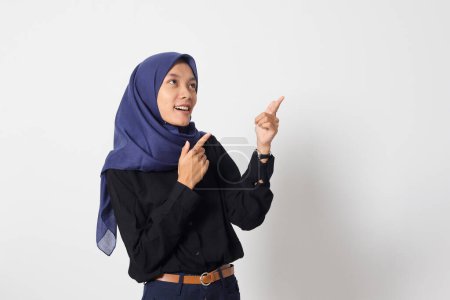 Téléchargez les photos : Portrait de femme hijab asiatique excitée en chemise décontractée faisant la promotion du produit, pointant du doigt vers le côté. Concept publicitaire. Image isolée sur fond blanc - en image libre de droit