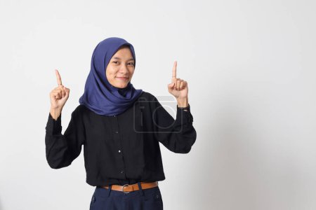 Téléchargez les photos : Portrait de femme asiatique excitée hijab en chemise décontractée promotion produit, pointant du doigt vers le haut. Concept publicitaire. Image isolée sur fond blanc - en image libre de droit
