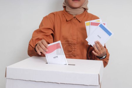 Foto de Primer plano de la mujer hiyab asiática insertando y poniendo el papel de votación en las urnas. Elecciones generales o Pemilu para el presidente y el gobierno de Indonesia. Imagen aislada sobre fondo blanco - Imagen libre de derechos