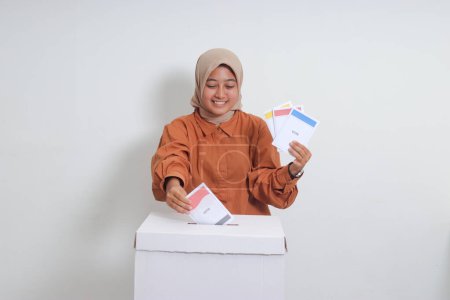 Foto de Retrato de una mujer hiyab asiática insertando y poniendo el papel de votación en las urnas. Elecciones generales o Pemilu para el presidente y el gobierno de Indonesia. Imagen aislada sobre fondo blanco - Imagen libre de derechos