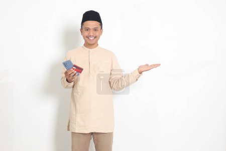 Téléchargez les photos : Portrait d'homme musulman asiatique attrayant en chemise koko avec un bonnet de crâne tenant un téléphone portable et une carte de crédit tout en pointant du doigt sur le côté. Image isolée sur fond blanc - en image libre de droit