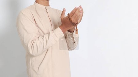 Close up portrait of religious Asian muslim man in koko shirt with skullcap praying earnestly with his hands raised, holding islamic beads. Devout faith concept. Isolated image on white background