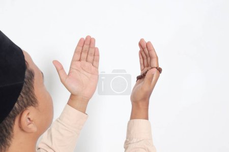Vista posterior retrato del hombre musulmán asiático religioso en camisa koko con gorra de calavera rezando fervientemente con las manos levantadas, sosteniendo cuentas islámicas. Concepto de fe devota. Imagen aislada sobre fondo blanco